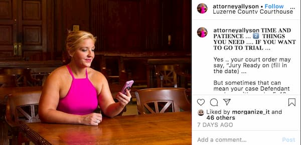 Allyson wearing a bright pink sleeveless dress sits behind a courtroom bench and holds her phone in her hand. 