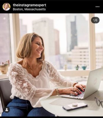 An Instagram post that shows Sue B Zimmerman sitting at her table in a white blouse and blue skirt as she smiles confidently at her computer.