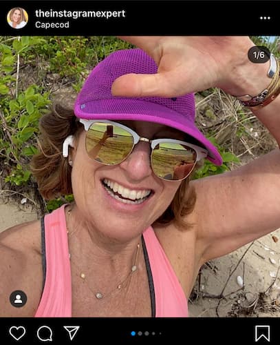 Sue B Zimmerman wears a purple baseball cap and pink tank top as she smiles and waves in her selfie.